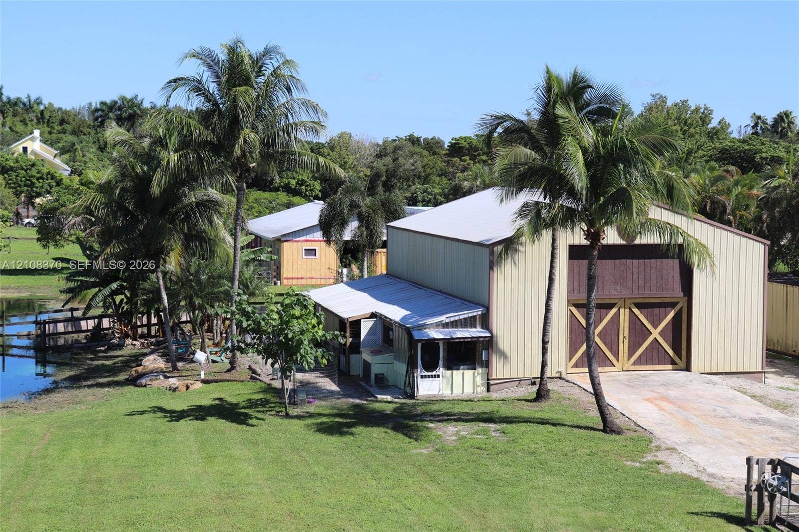 18901 SW 63rd Street, Southwest Ranches, FL 33332 Metal-sided workshop featuring large carriage-style doors, a concrete driveway, and mature palm tree landscaping on a waterfront lot