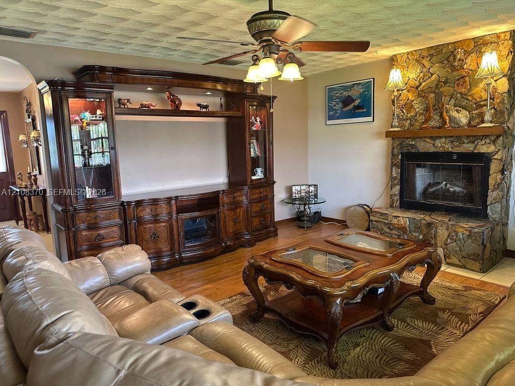 18901 SW 63rd Street, Southwest Ranches, FL 33332 Living area featuring a floor-to-ceiling natural stone fireplace with a raised hearth, wood-finish flooring, built-in wooden media cabinetry, and a ceiling fan with integrated lighting