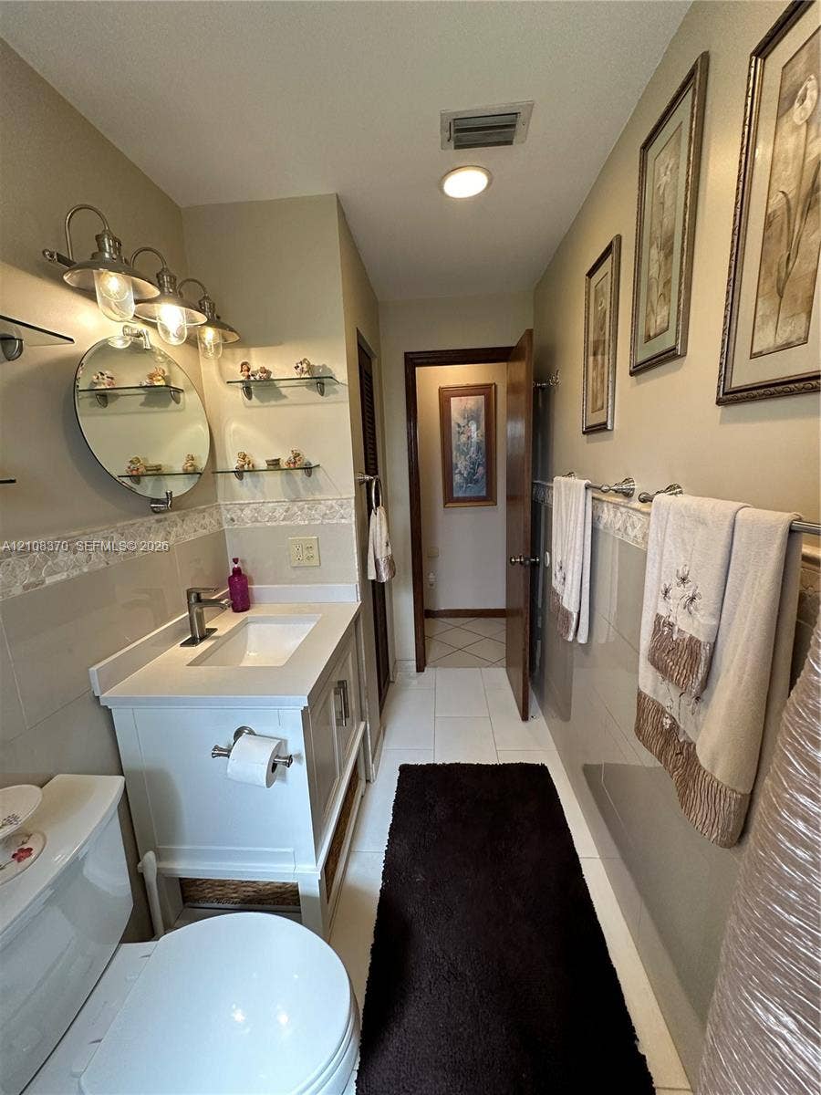 18901 SW 63rd Street, Southwest Ranches, FL 33332 White vanity with integrated basin, polished metal hardware, circular wall-mounted mirror, glass shelving, and tile wainscoting