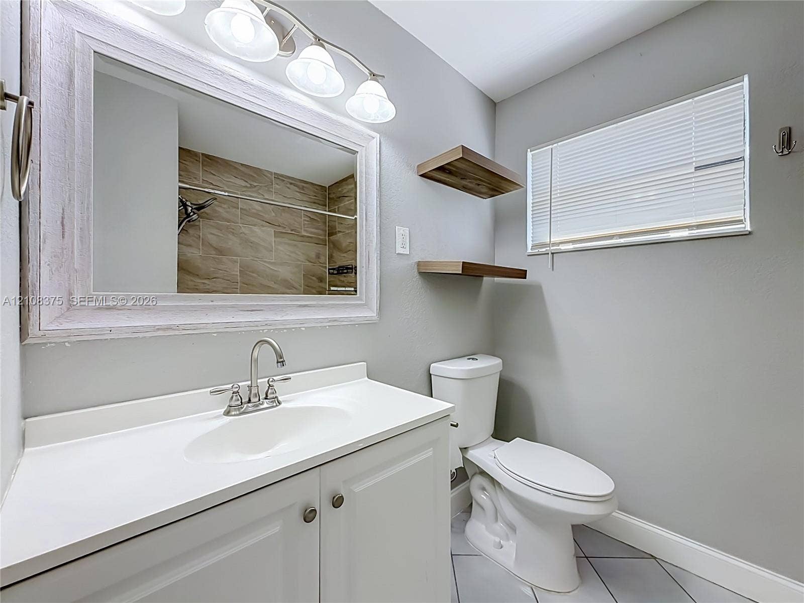 3330 NE 14th Terrace, Pompano Beach, FL 33064 Bathroom featuring a white vanity cabinet with a solid-surface countertop, an integrated sink, a framed mirror with a four-light fixture, wood-finish floating shelves, and tile flooring