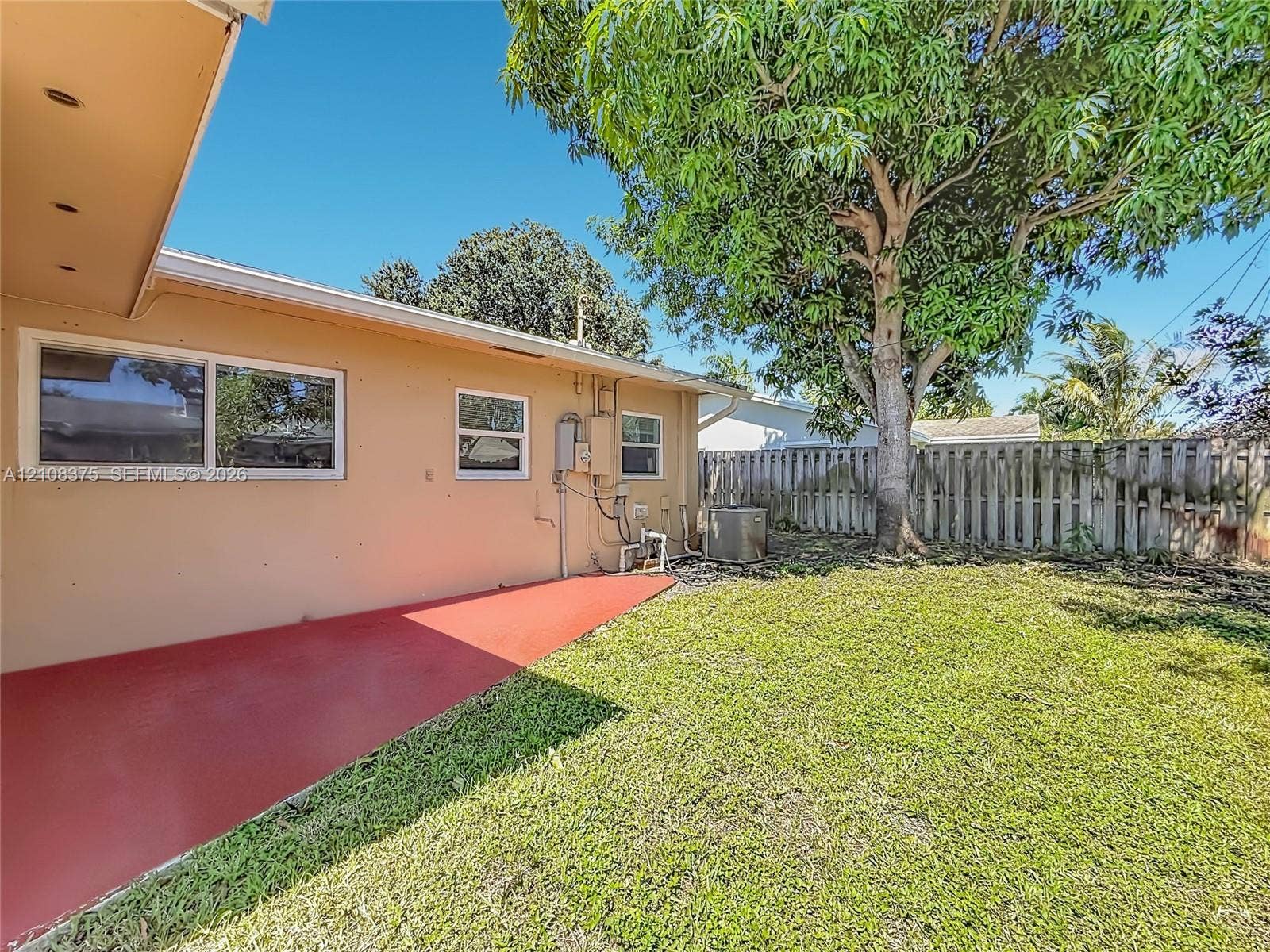 3330 NE 14th Terrace, Pompano Beach, FL 33064 Rear exterior featuring a concrete patio, grassy yard, mature tree, and wood-panel privacy fencing