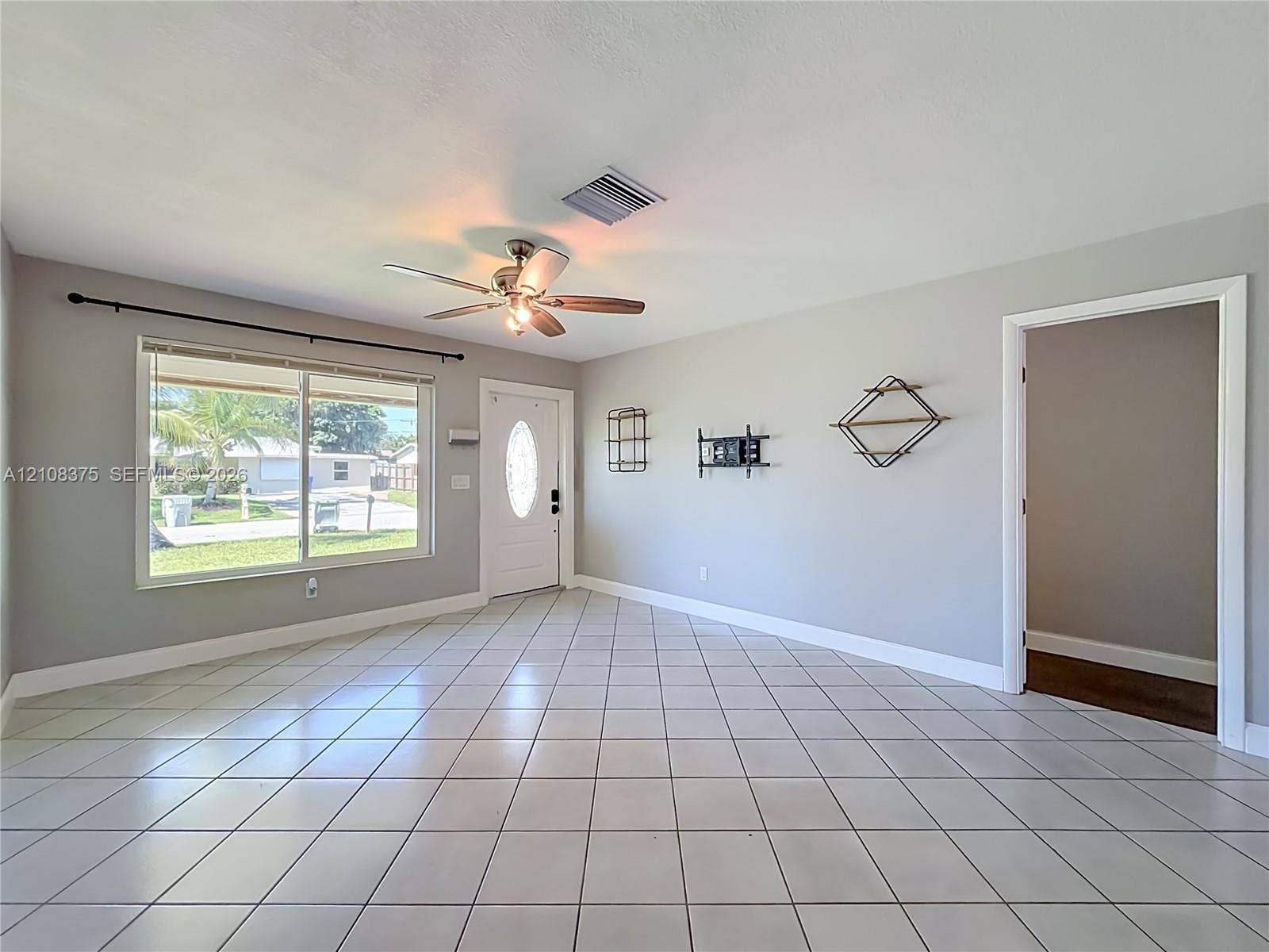 3330 NE 14th Terrace, Pompano Beach, FL 33064 Neutral-toned interior featuring large-format ceramic tile flooring, a multi-pane window with hardware, a solid-panel front door with an oval glass insert, and a ceiling-mounted fan with integrated lighting