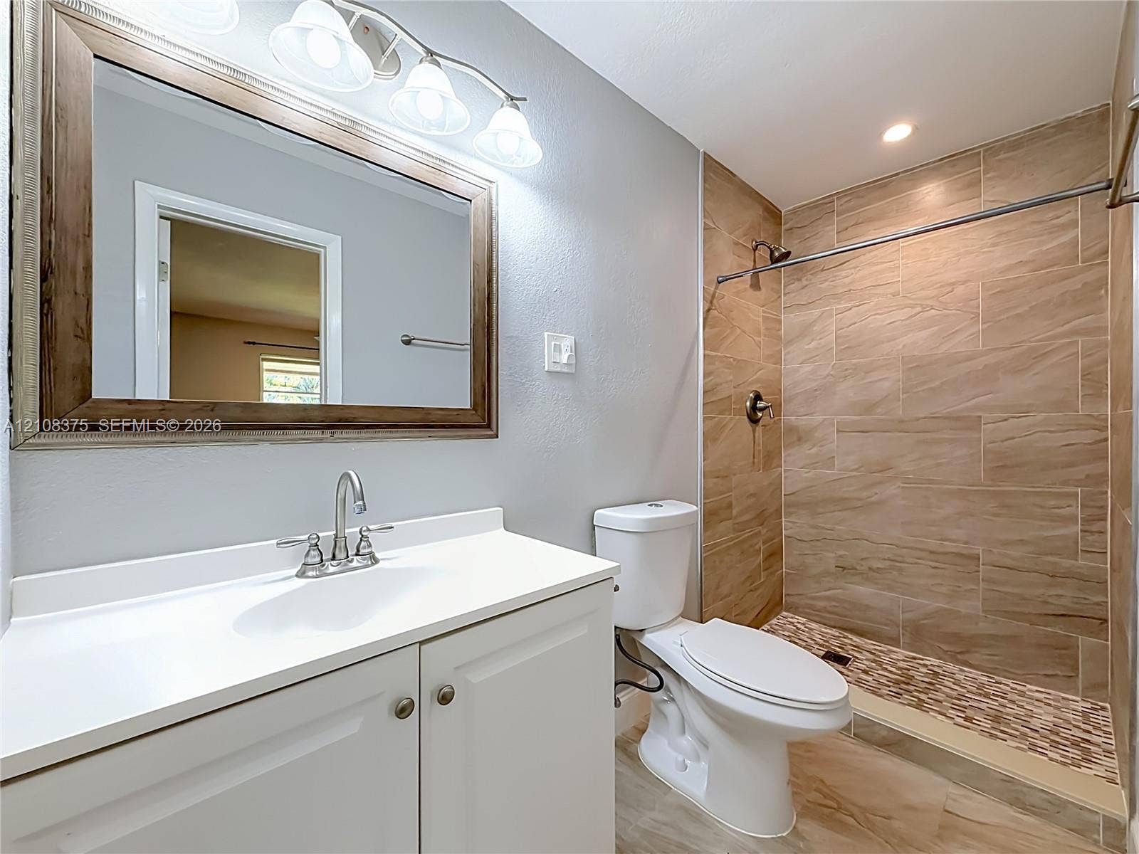 3330 NE 14th Terrace, Pompano Beach, FL 33064 Bathroom featuring a vanity with a white countertop and sink, walk-in shower with horizontal tile surround, mosaic stone-finish shower floor, and a three-light wall-mounted fixture