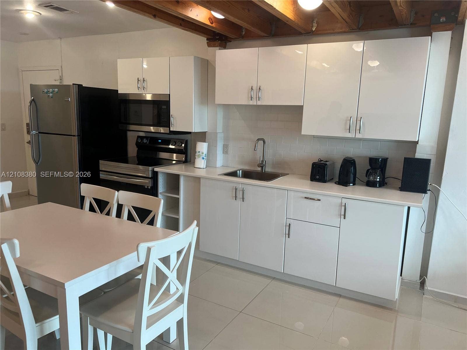 5445 Collins Avenue, #BAY11, Miami Beach, FL 33140 Kitchen featuring high-gloss white cabinetry, light-toned solid surface countertops, a white subway tile backsplash, stainless steel appliances, and exposed wood ceiling beams