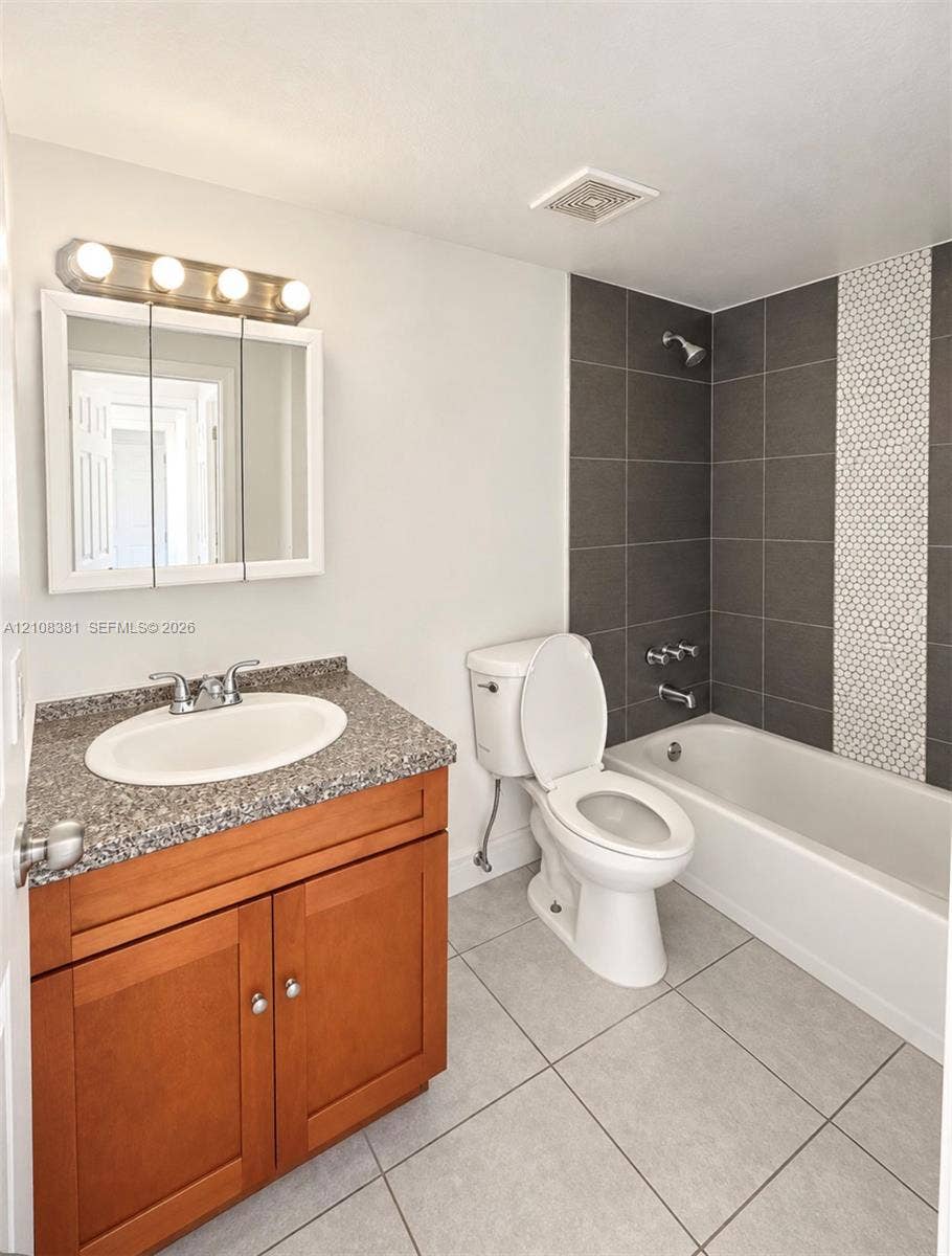 621 NW 177th Street, #101, Miami Gardens, Miami Gardens, FL 33169 Bathroom featuring a wood-finish vanity with a stone-composite countertop, integrated medicine cabinet, tile-surround tub with a decorative mosaic accent strip, and large-format floor tiling