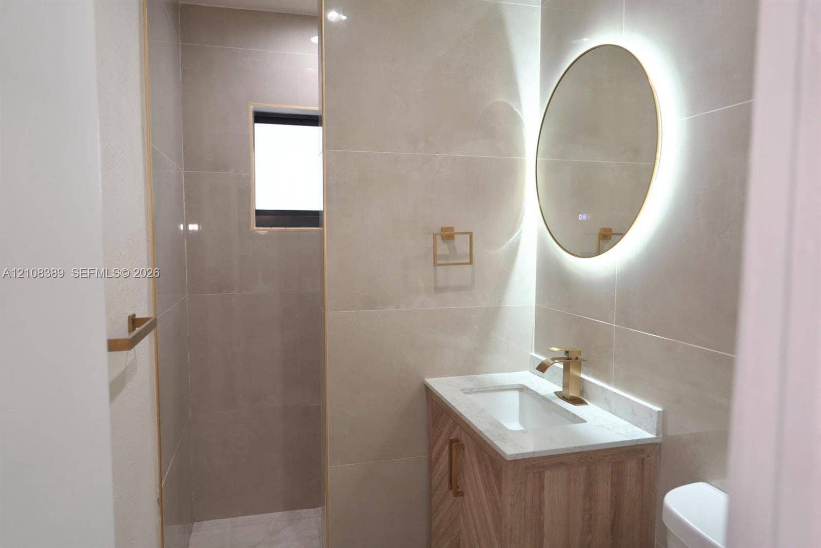 Wood-finish vanity with a stone countertop, wall-mounted circular backlit mirror, brushed gold-tone hardware, and floor-to-ceiling tile work