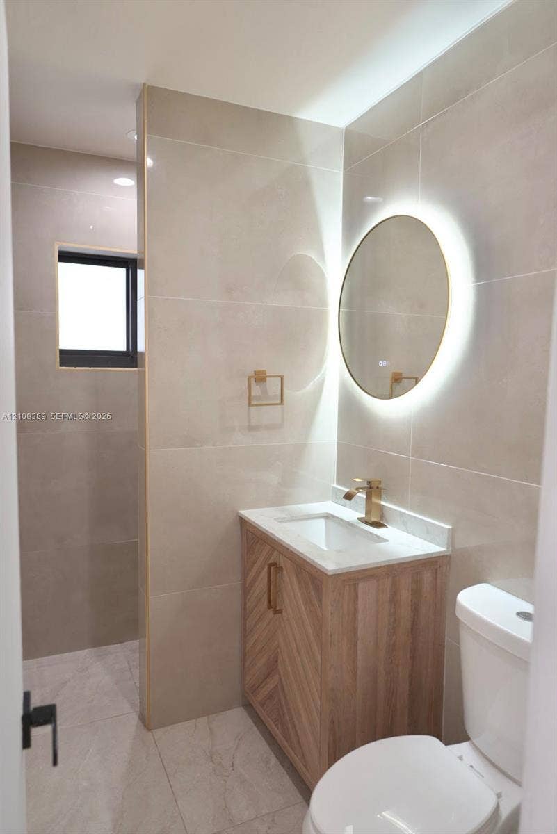 Bathroom featuring large-format wall tile, wood-finish vanity with chevron detailing, stone-surface countertop, gold-tone hardware, and circular back-lit mirror