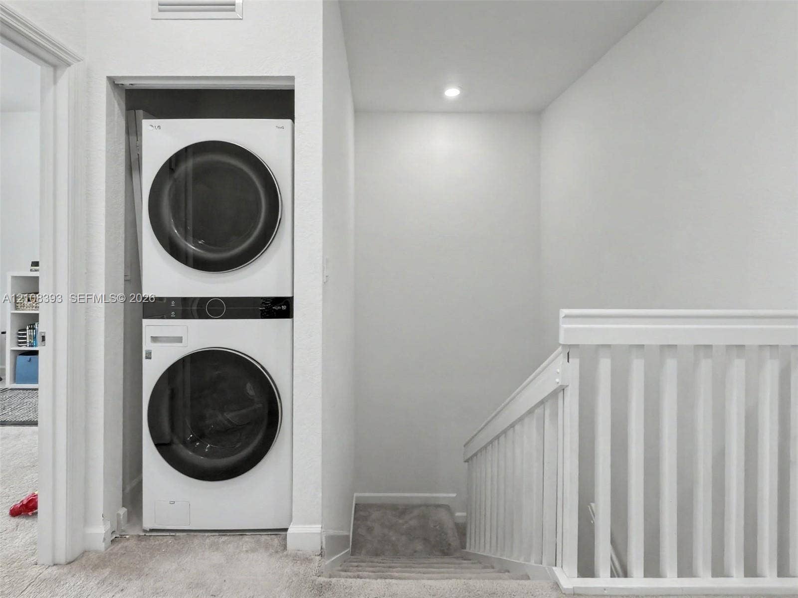 Stacked laundry unit with front-loading appliances, recessed ceiling lighting, and white wooden stair railing