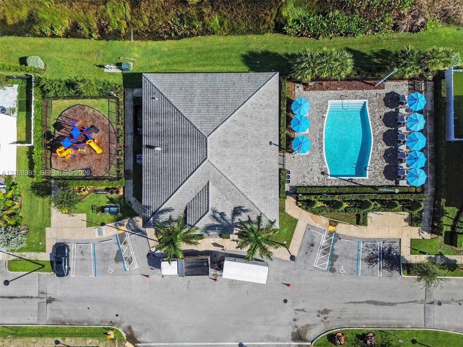 Aerial perspective of a community facility featuring a swimming pool, paved pool deck, enclosed recreation area, and paved parking area