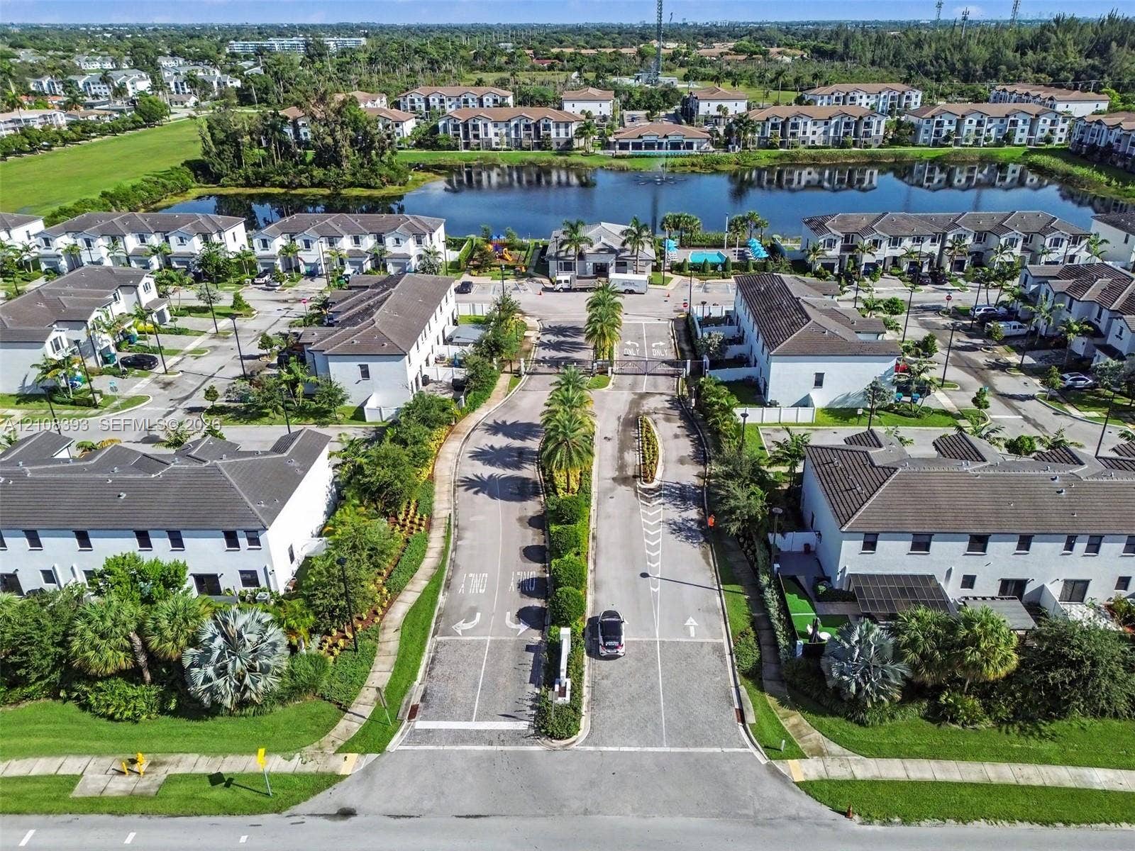 Aerial view of a gated community featuring multi-unit residential structures, paved roadways, manicured landscaping with palm trees, and a central pond