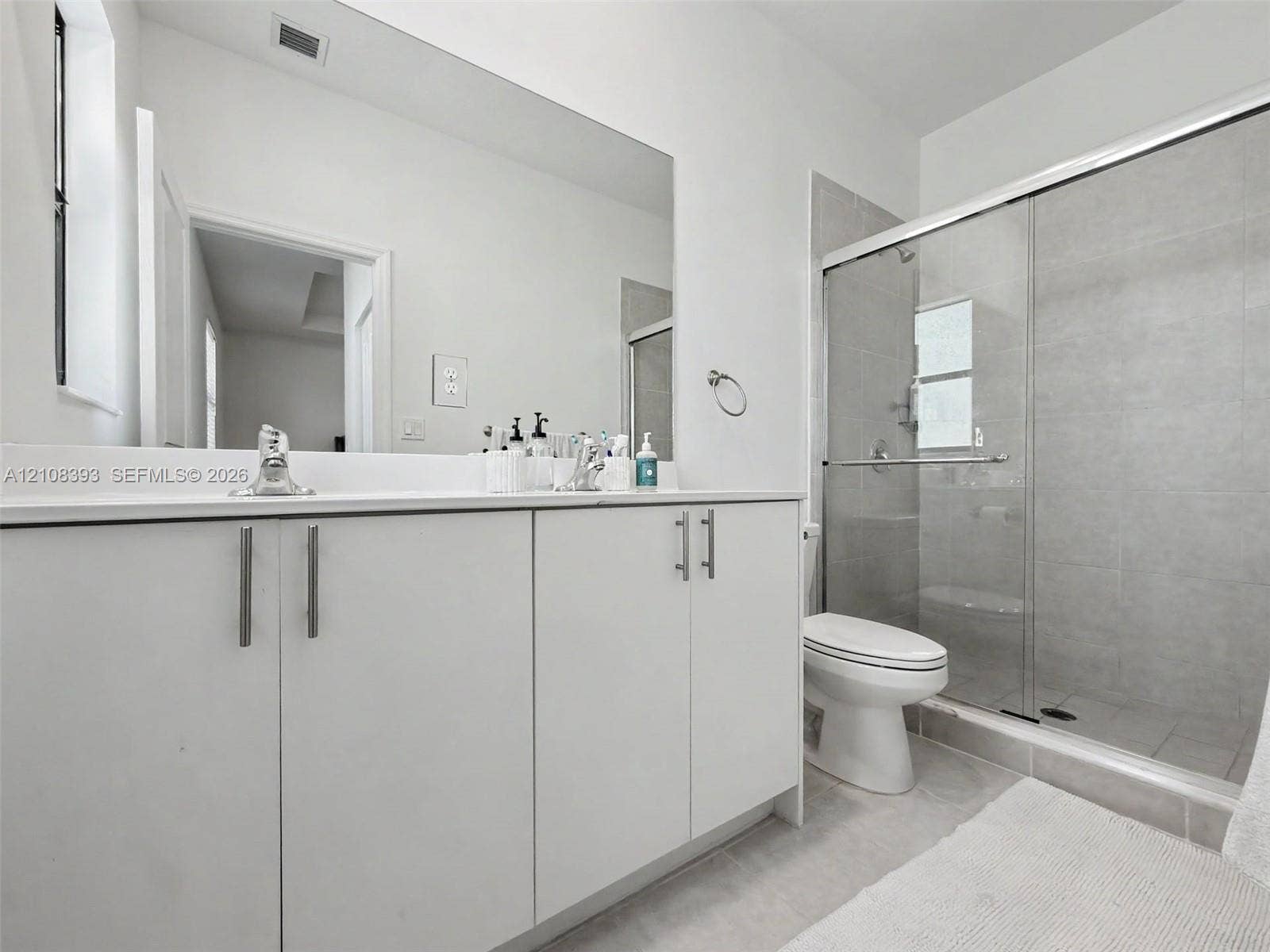 Bathroom featuring white shaker-style cabinetry, solid-surface countertop, frameless glass shower enclosure with neutral-toned wall tile, and wall-to-wall vanity mirror