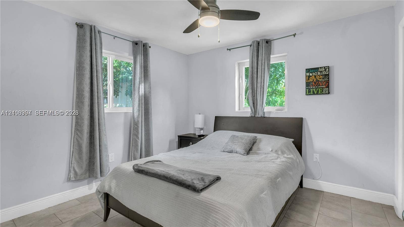 Neutral-toned bedroom featuring large format tile flooring, light-colored walls, dual windows, and a central ceiling fan with integrated lighting