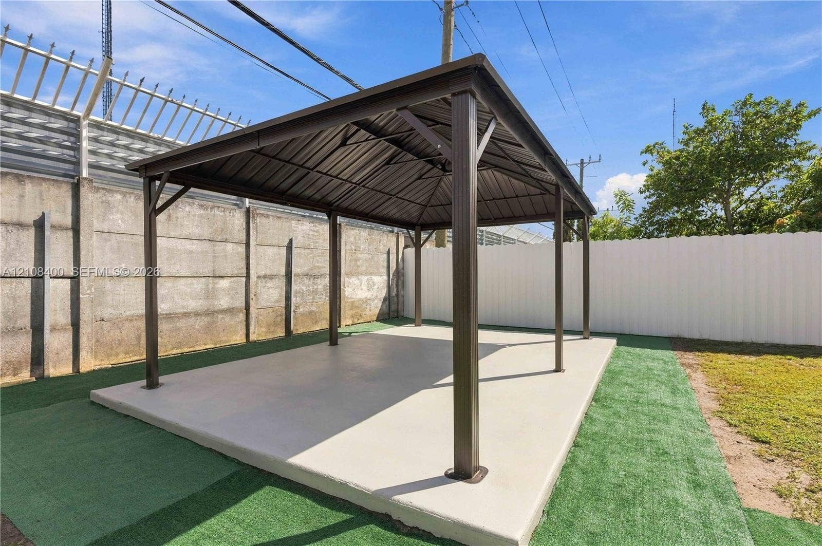 2551 SW 53rd Avenue, West Park, West Park, FL 33023 Outdoor gazebo featuring a dark metal frame, a peaked metal roof, and a concrete slab foundation, bordered by a concrete perimeter wall and synthetic turf