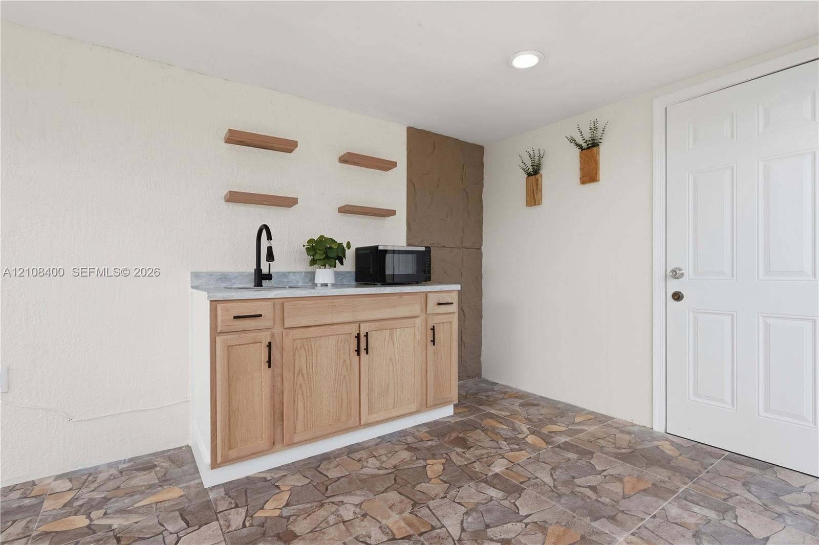 2551 SW 53rd Avenue, West Park, West Park, FL 33023 Custom cabinetry with light wood-finish doors, stone-pattern countertop, matte black faucet, floating wall shelving, and flagstone-style flooring