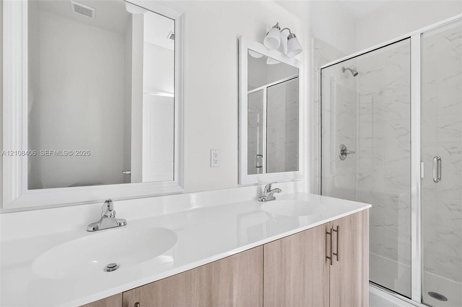 15650 SW 136th Street, #212, Miami, FL 33196 Dual-sink vanity with a white countertop and wood-finish cabinetry, framed mirrors, and a glass-enclosed shower with light-toned wall tile