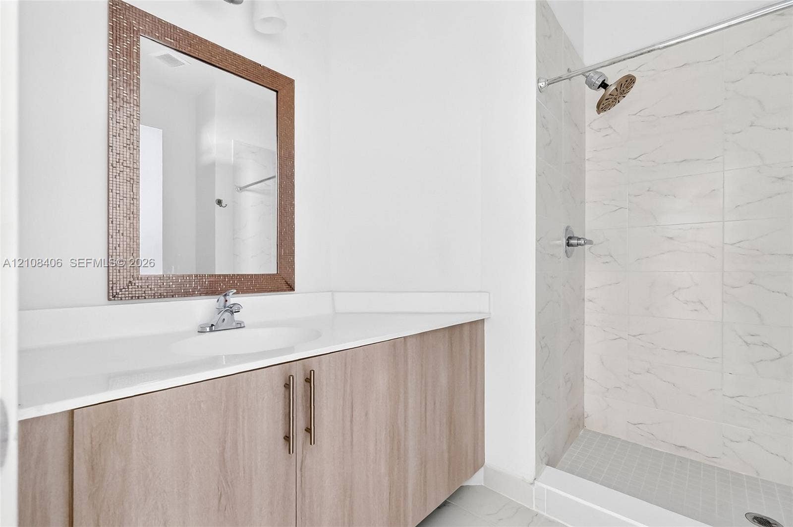 15650 SW 136th Street, #212, Miami, FL 33196 Bathroom vanity featuring a wood-finish cabinet, white solid-surface countertop, and rectangular mirror with a mosaic frame, alongside a walk-in shower with light-toned wall tile and a silver-finish shower head
