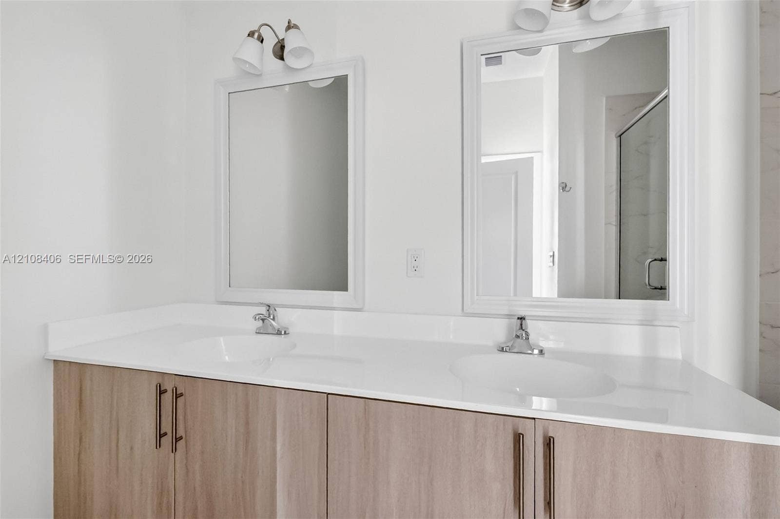 15650 SW 136th Street, #212, Miami, FL 33196 Dual-sink vanity with a white solid-surface countertop, light wood-finish cabinetry, brushed metal hardware, and framed mirrors with overhead light fixtures