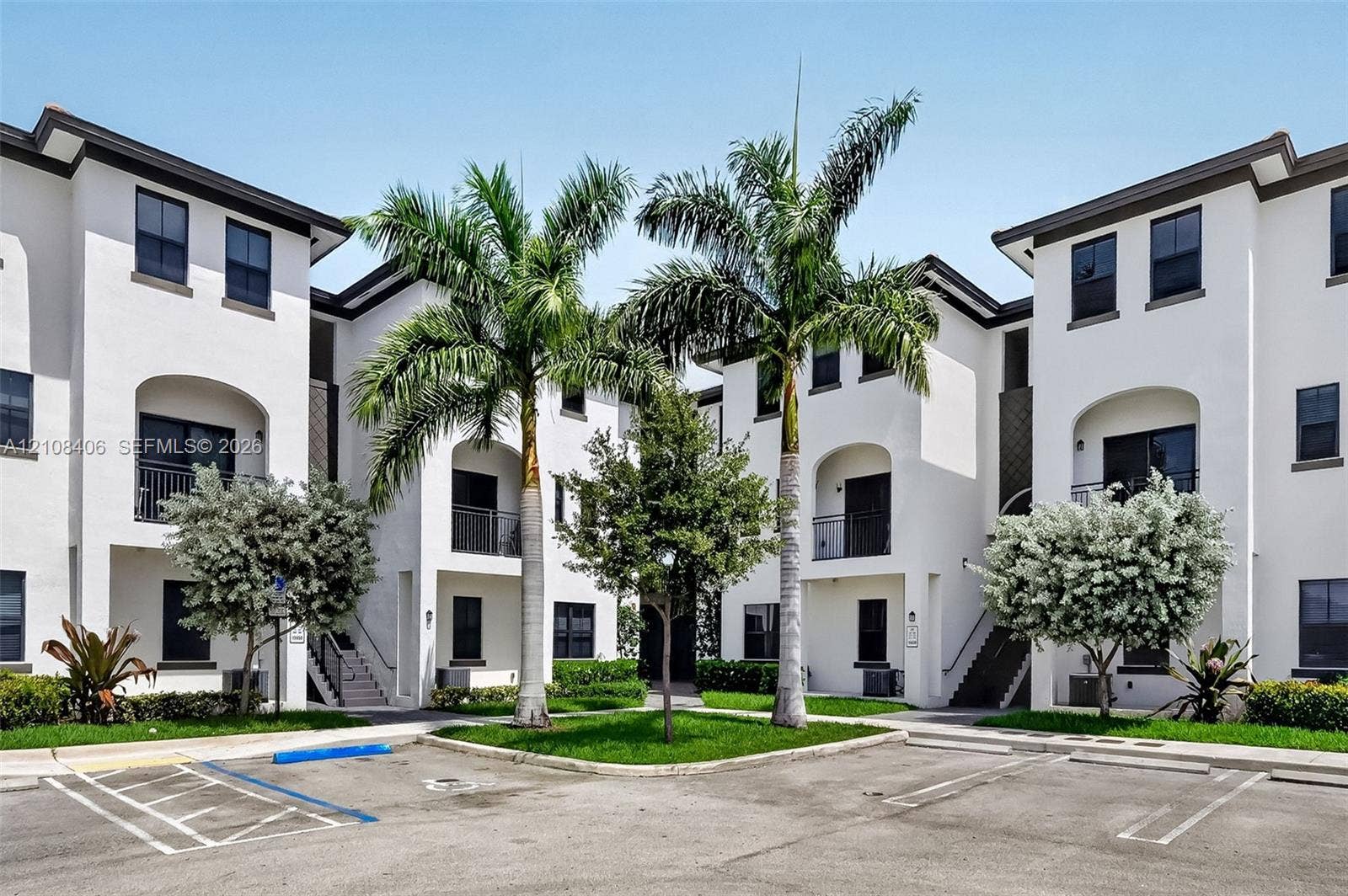 15650 SW 136th Street, #212, Miami, FL 33196 Stucco exterior featuring arched entryways, balconies with metal railings, and multi-level design, complemented by a landscaped courtyard with palm trees and manicured lawns