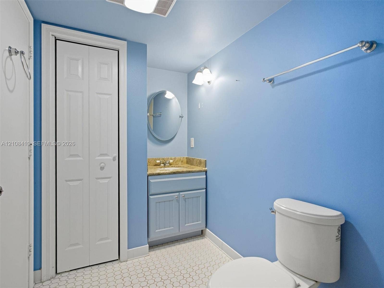 7556 Stirling Road, #124, Davie, Davie, FL 33024 Bathroom featuring a single vanity with stone-style countertop, oval wall-mounted mirror, white ceramic tile flooring, bifold closet doors, and brushed metallic hardware