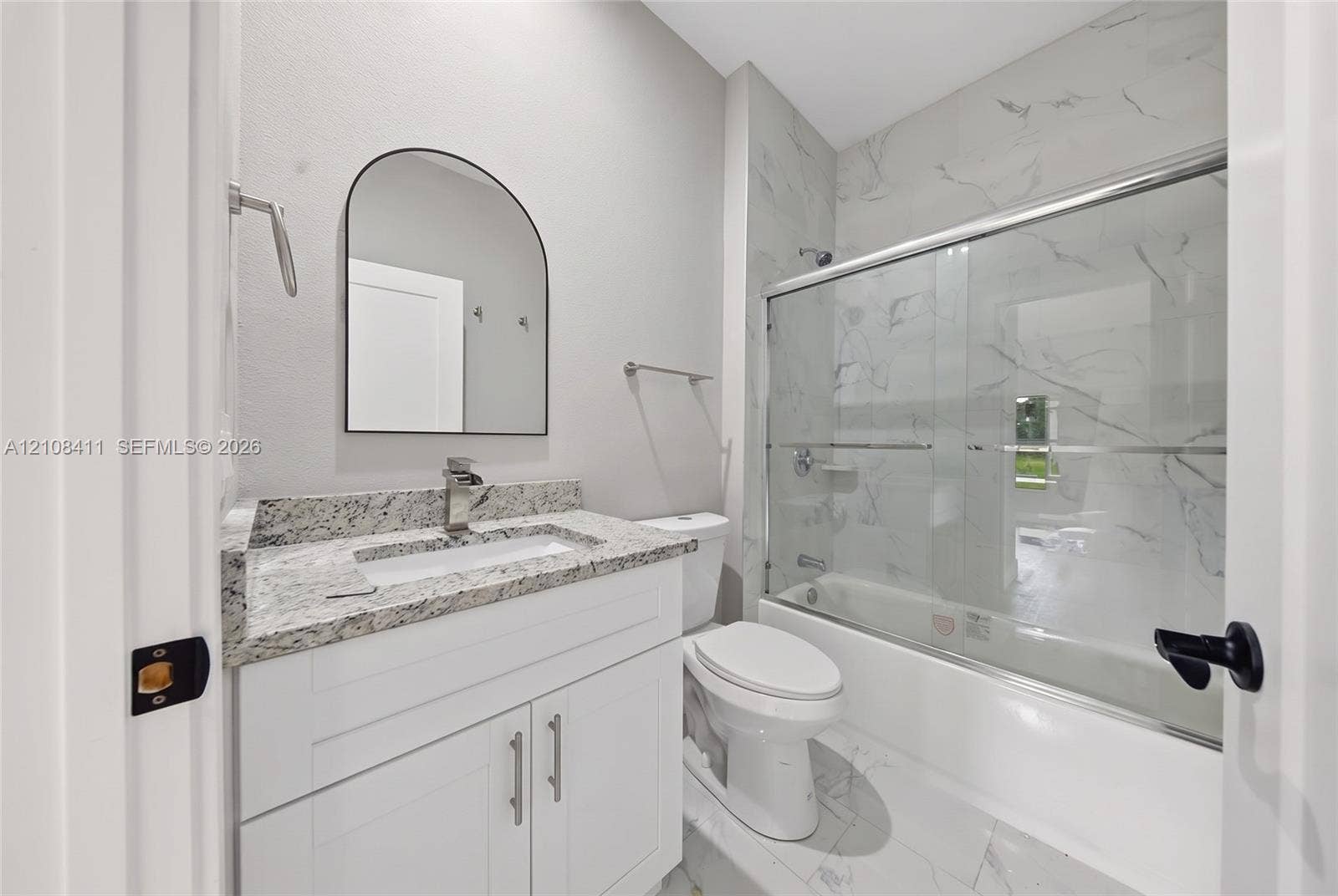 527 Avenida Del Sur, Clewiston, FL 33440 Bathroom featuring a white shaker-style vanity, stone-finish countertop, arched wall-mounted mirror, and a shower-tub combination with full-height tile surround