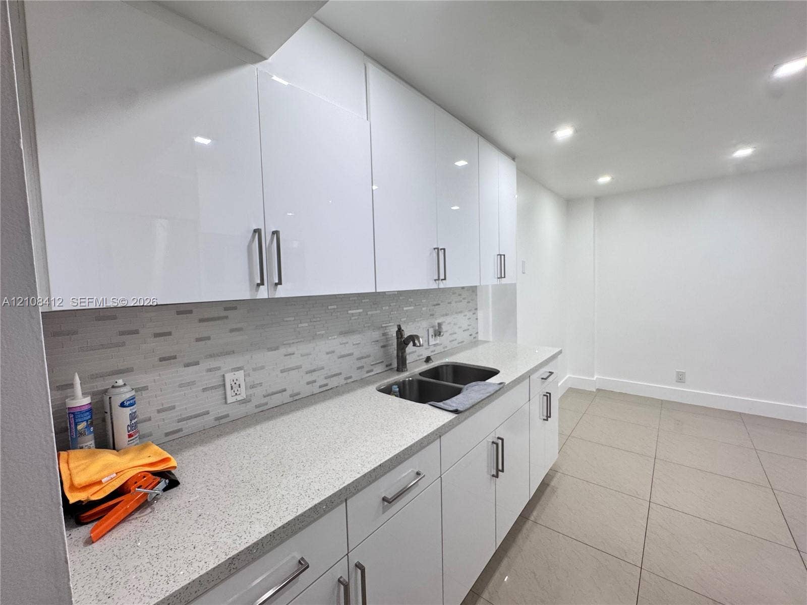 Glossy white cabinetry, stone-finish countertops, linear tile backsplash, stainless steel dual-basin sink with a gooseneck faucet, and recessed ceiling lighting