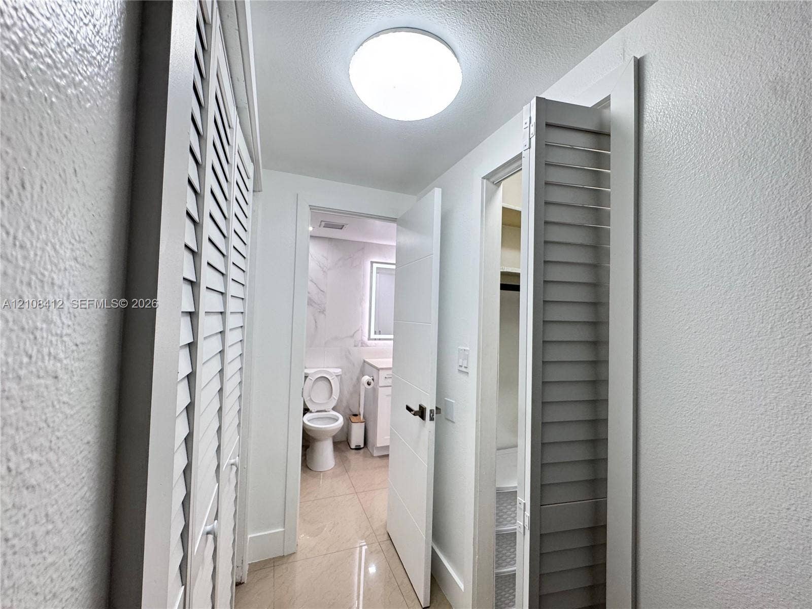 Hallway featuring dual sets of white louvered doors, high-gloss large-format tile flooring, a minimalist solid-core interior door, and built-in closet shelving