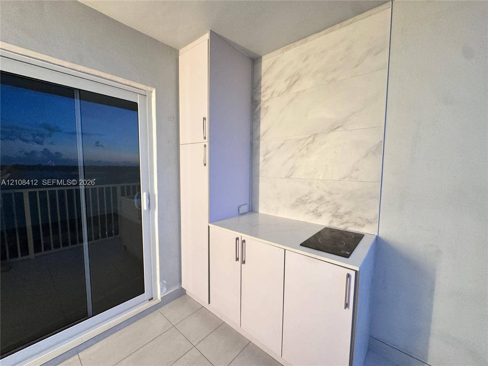 Built-in cabinetry featuring white flat-panel doors with metallic hardware, stone-finish backsplash, smooth-surface electric cooktop, and large format floor tile