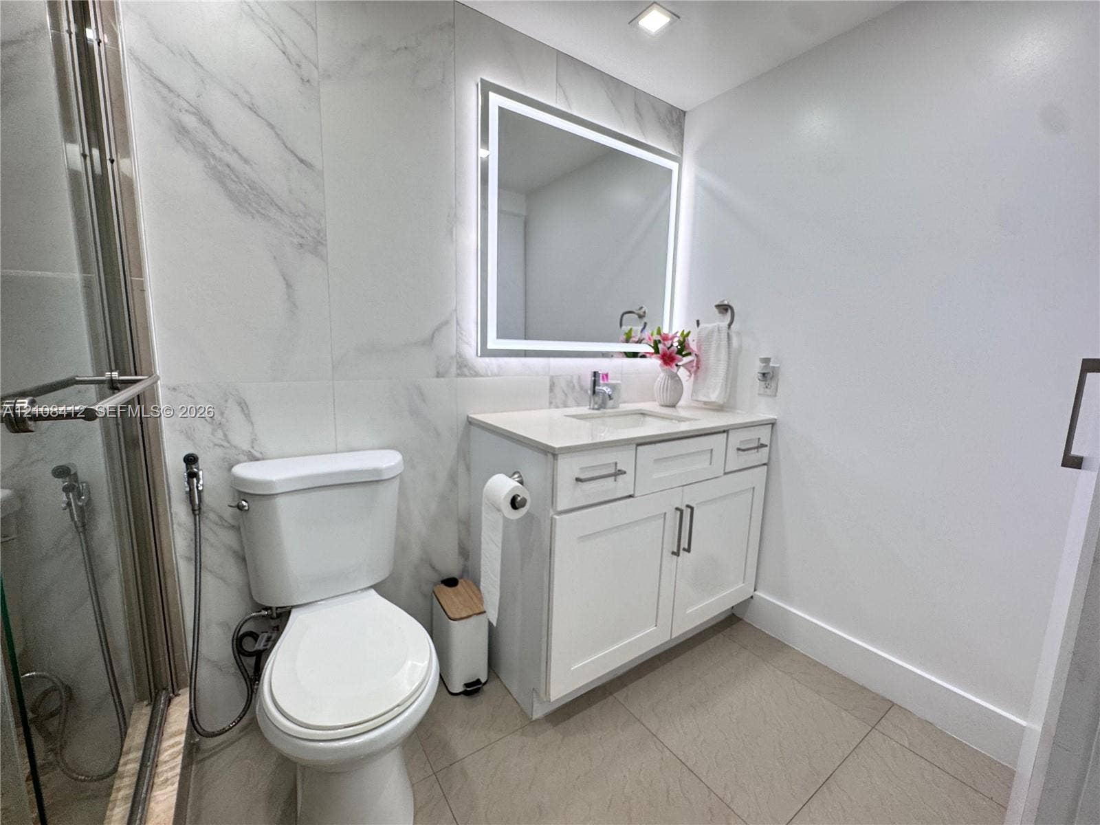 Bathroom featuring a white vanity cabinet with a light-toned countertop, an illuminated mirror, large-format wall tiling with a veined pattern, and a glass-enclosed shower
