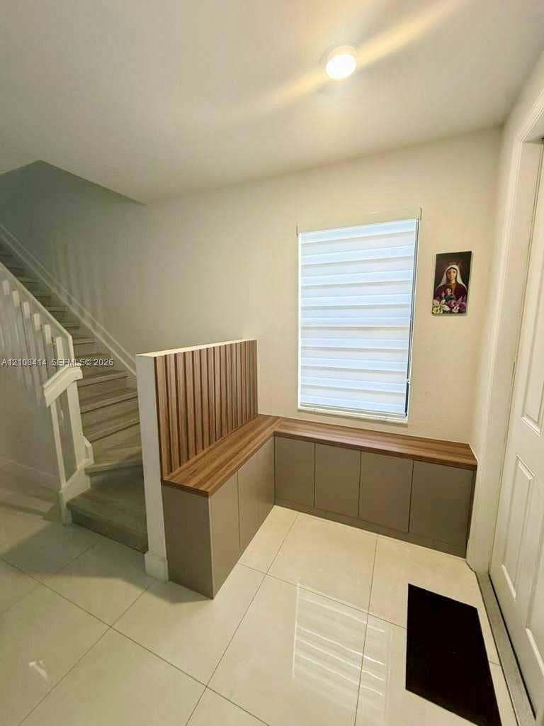 Built-in L-shaped cabinetry featuring wood-finish countertops and vertical slat detailing, paired with large-format tile flooring and a window equipped with dual-layered adjustable shades
