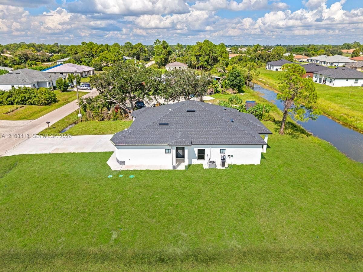 2235 Wayne Avenue S, Lehigh Acres, FL 33973 Single-story residence featuring a shingled roof, white exterior finish, concrete driveway, and expansive lawn area situated along a canal
