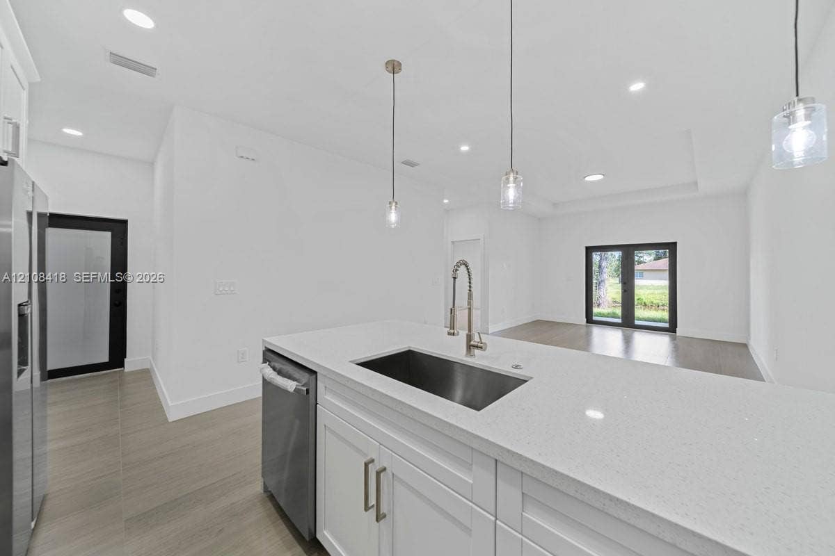2235 Wayne Avenue S, Lehigh Acres, FL 33973 Kitchen island featuring white shaker-style cabinetry, quartz countertops, an undermount stainless steel sink, and a gooseneck faucet, accented by glass pendant lighting and light-toned wood-finish flooring