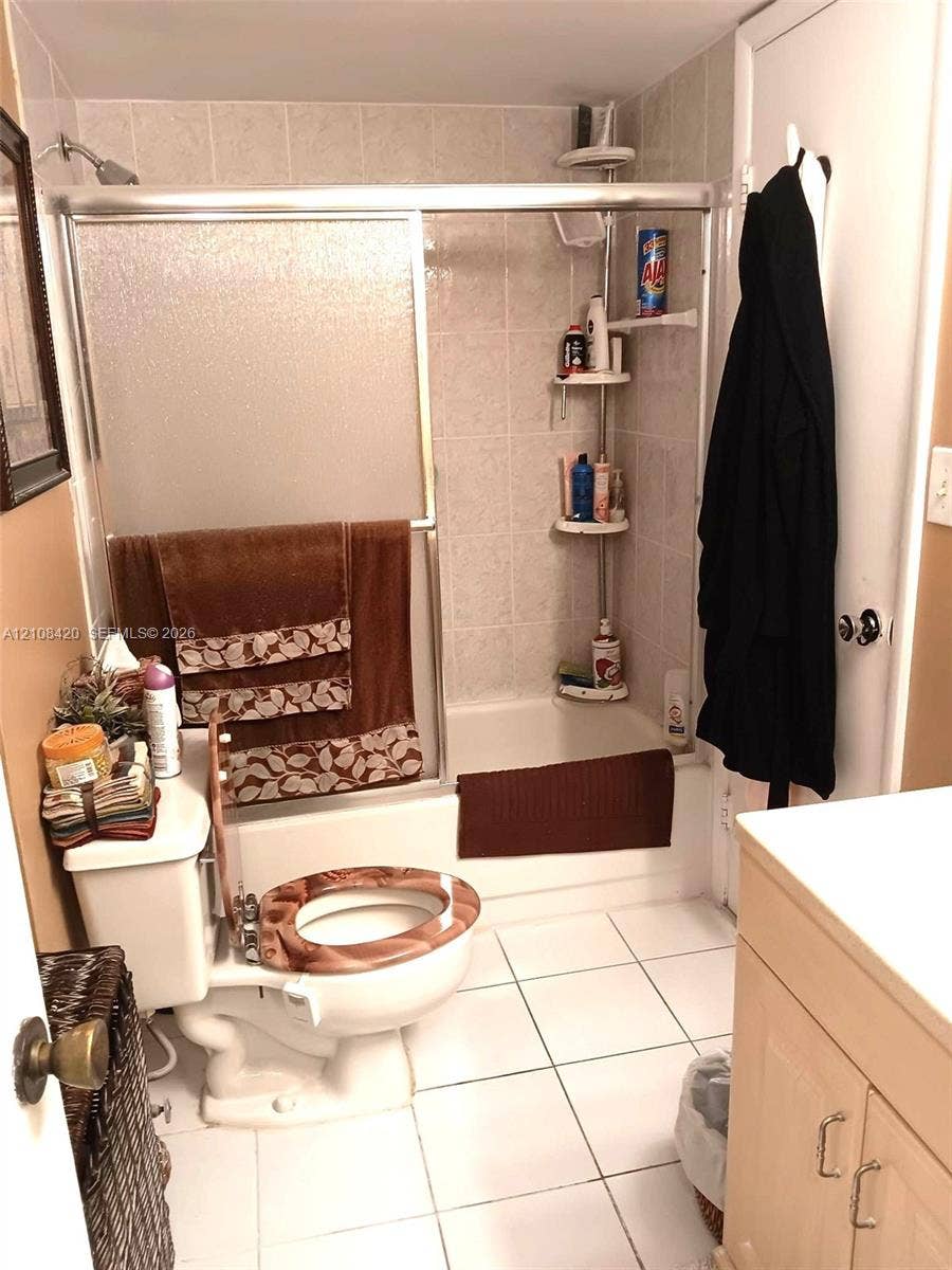 3545 NE 166th Street, #306, North Miami Beach, FL 33160 Bathroom featuring a tub-shower combination with a sliding glass door enclosure and light-toned tile surround, white square floor tiling, and wood-finish cabinetry