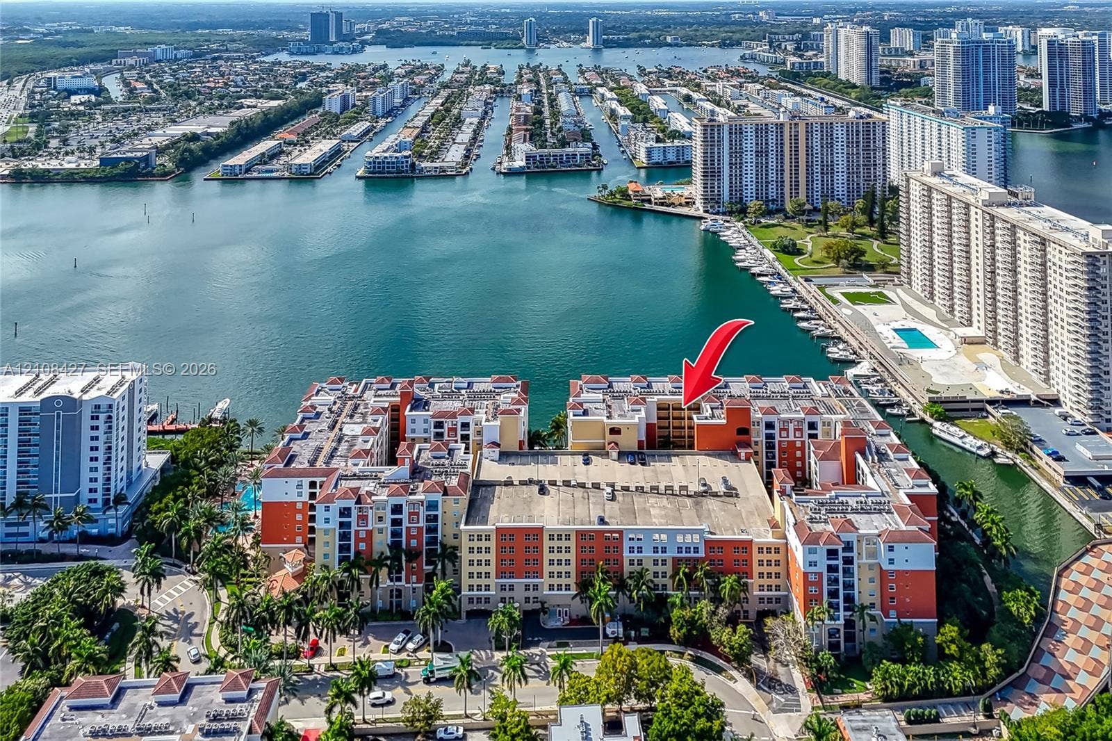 Aerial perspective of a multi-story residential complex situated on a waterfront peninsula, featuring a structured parking facility, colorful stucco facade, and multiple outdoor balconies