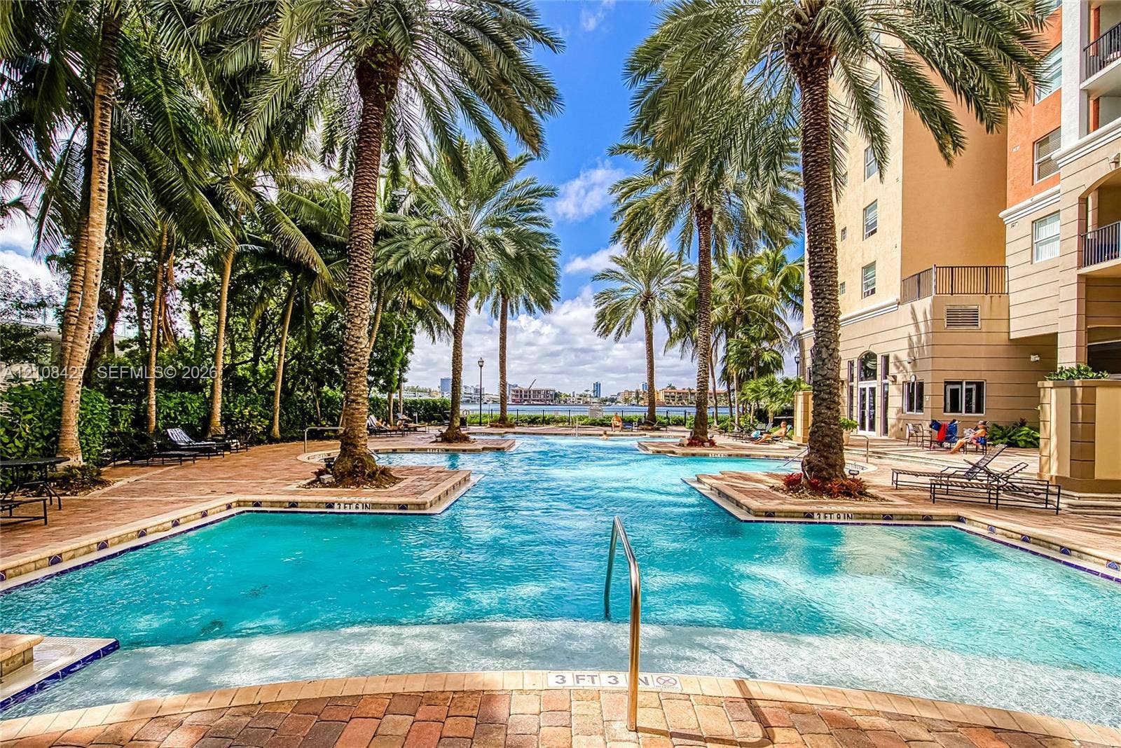 Resort-style swimming pool featuring brick paver decking, integrated palm tree landscaping, shallow-entry steps, and stainless steel handrails