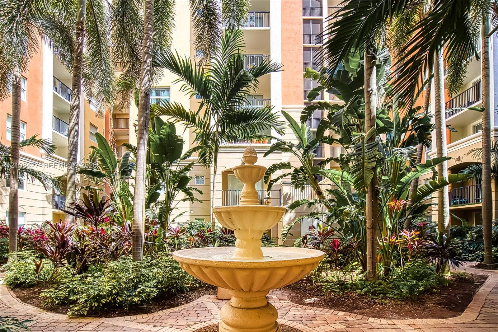 Central courtyard featuring a multi-tiered stone fountain, paved brick pathways, mature palm trees, and lush tropical landscaping