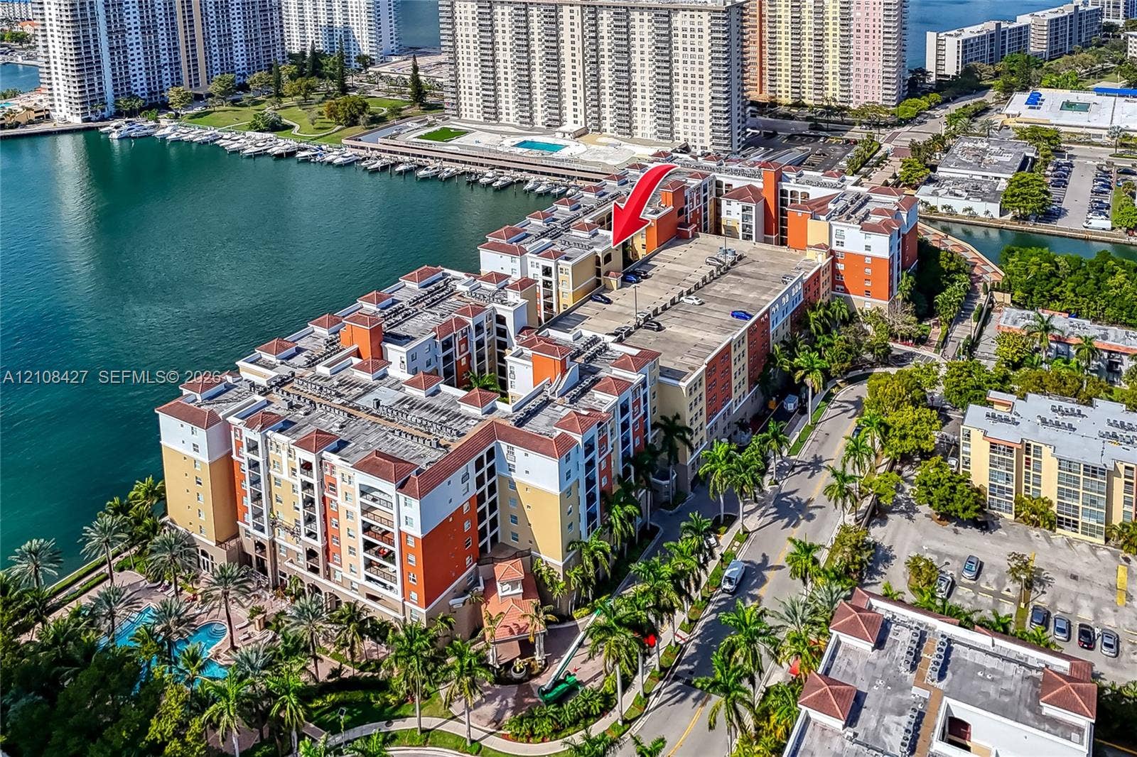 Aerial perspective of a multi-story waterfront complex featuring a swimming pool, mature palm trees, multi-tone masonry exterior, and tile roofing
