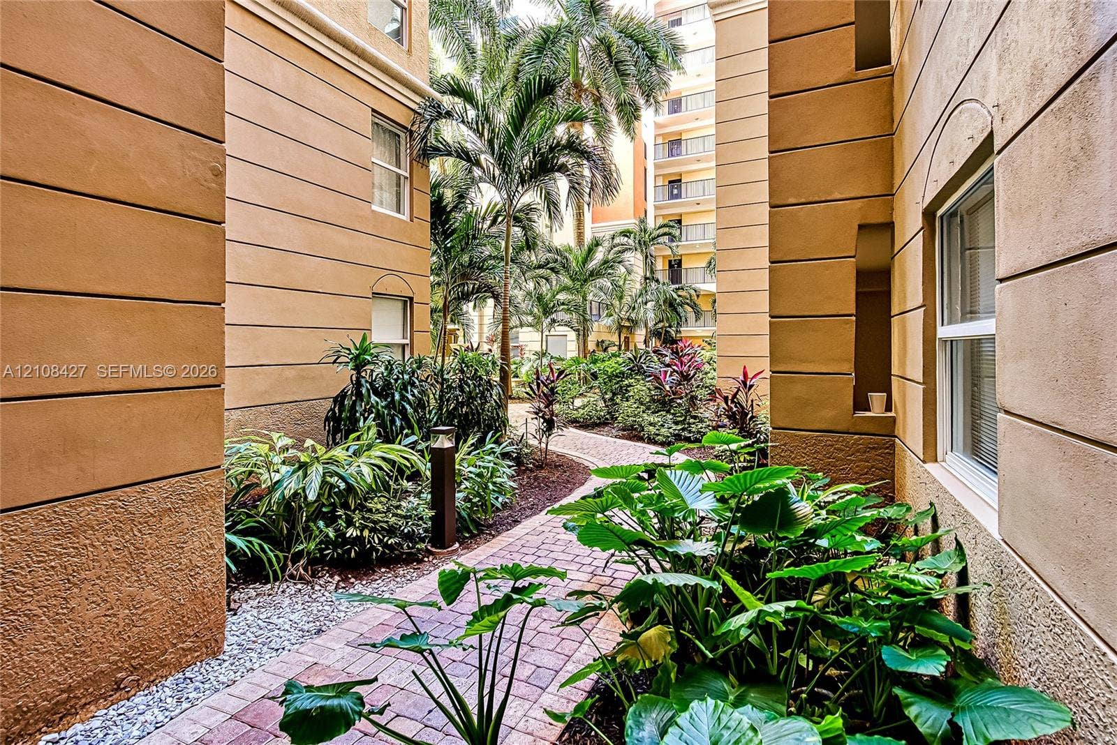 Stucco building exterior with recessed architectural detailing, paver walkway, and lush tropical landscaping