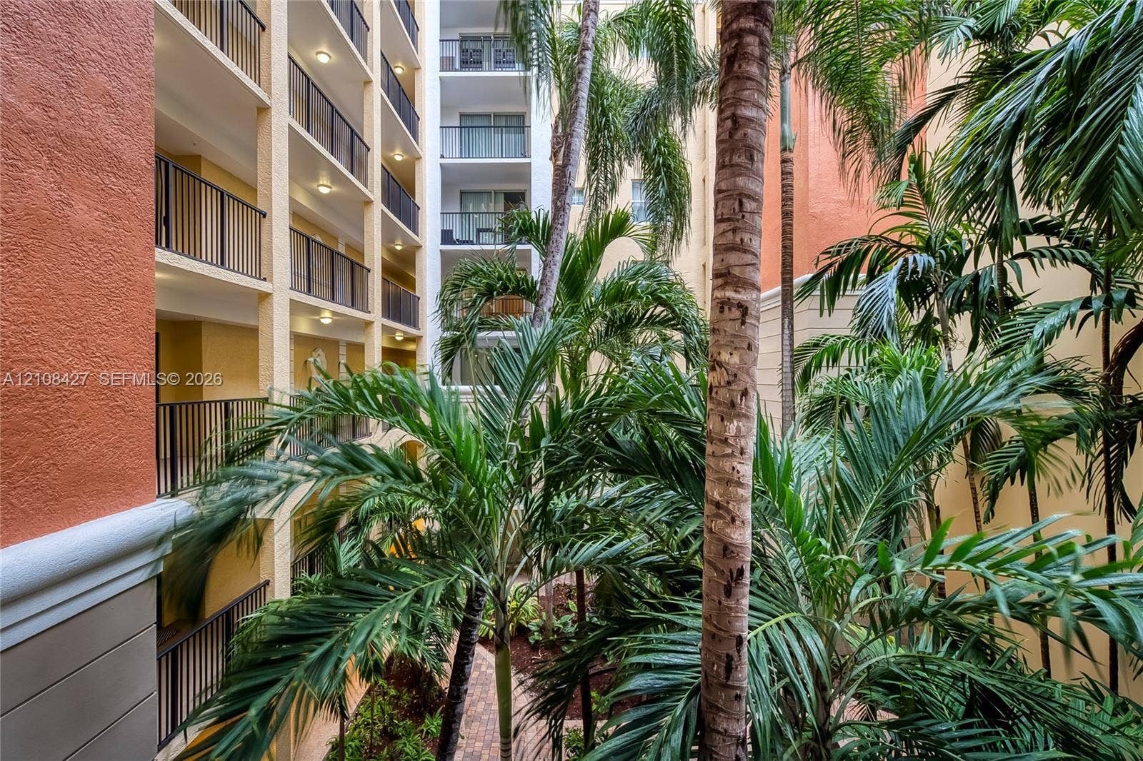 Multi-story atrium featuring private balconies with iron railings, a central courtyard with tropical palms, and brick paver walkways