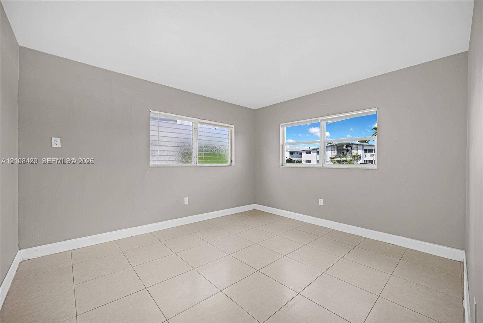 1430 NE 171st Street, North Miami Beach, FL 33162 Neutral-toned interior space featuring light-colored ceramic tile flooring, neutral wall paint, and dual windows with adjustable jalousie-style glass panes