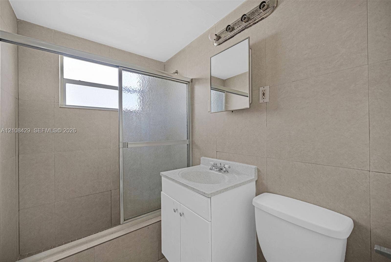 1430 NE 171st Street, North Miami Beach, FL 33162 Bathroom featuring floor-to-ceiling tile, single-basin vanity with a stone-finish countertop, sliding glass shower enclosure, and rectangular mirrored medicine cabinet