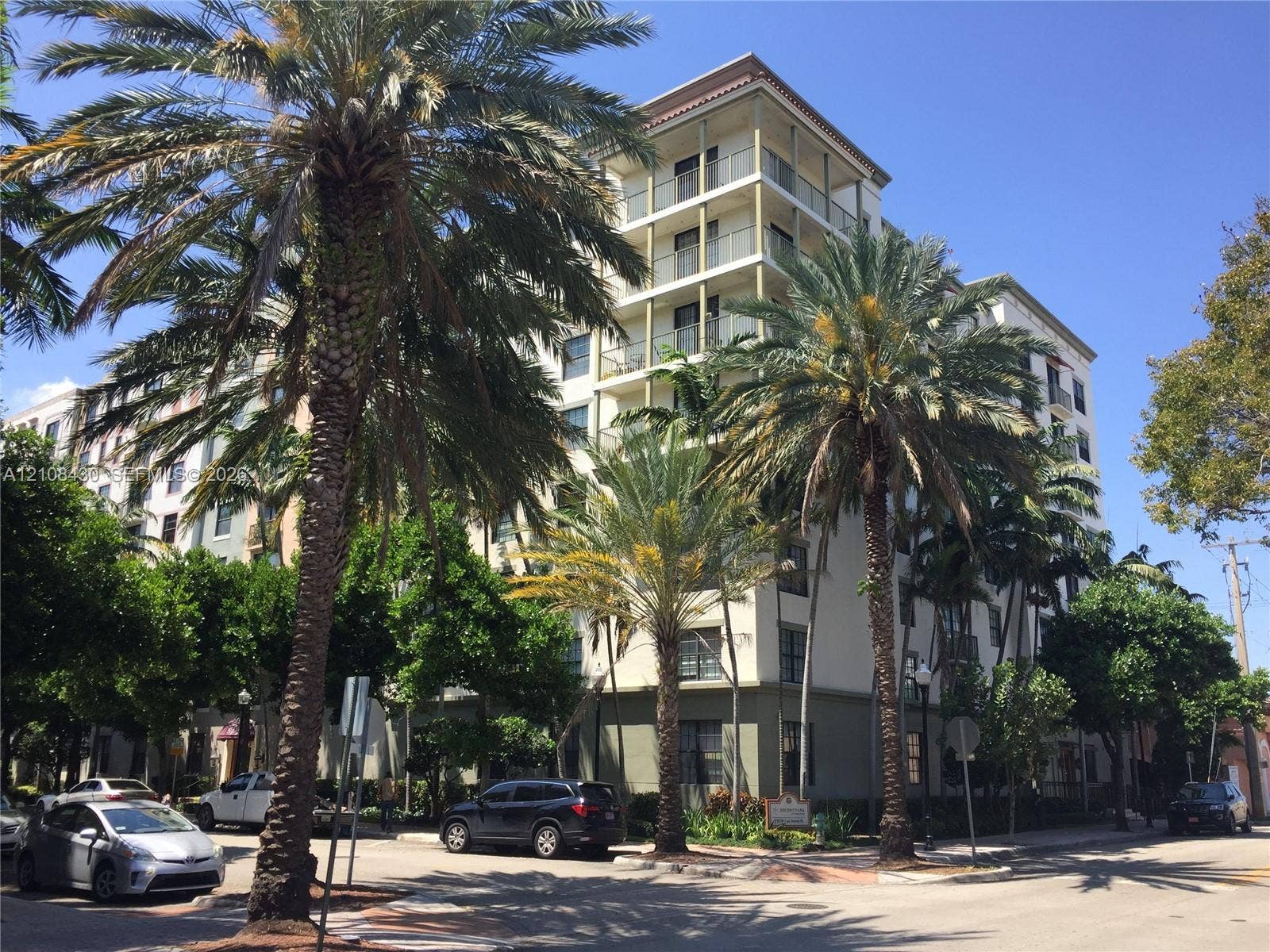 1919 Van Buren Street, #219A, Hollywood, FL 33020 Mid-rise multi-story building exterior featuring a stucco facade, tiered balconies with glass railings, a flat roofline, and mature palm tree landscaping