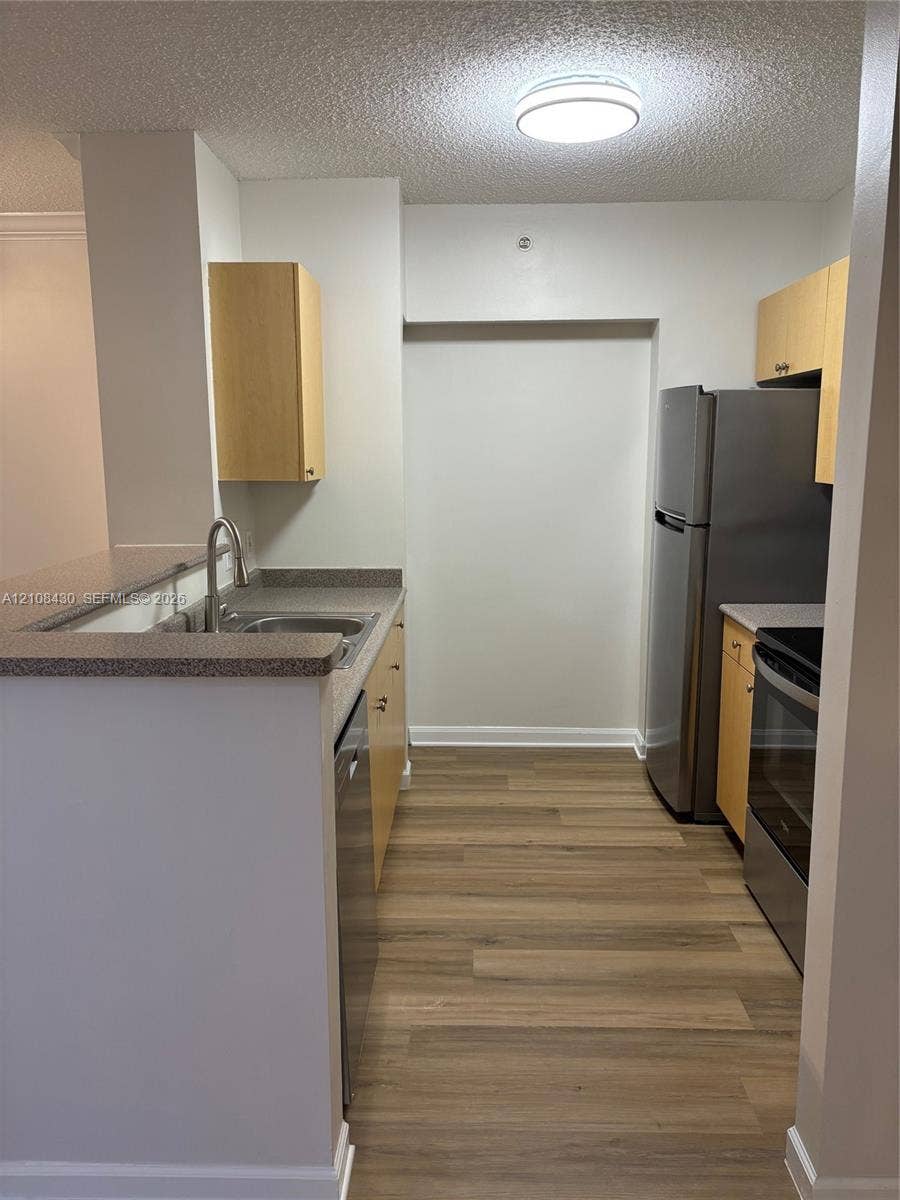 1919 Van Buren Street, #219A, Hollywood, FL 33020 Kitchen featuring wood-finish flooring, stainless steel appliances, light-toned wood cabinetry, stone-composite countertops, and an integrated double-basin sink