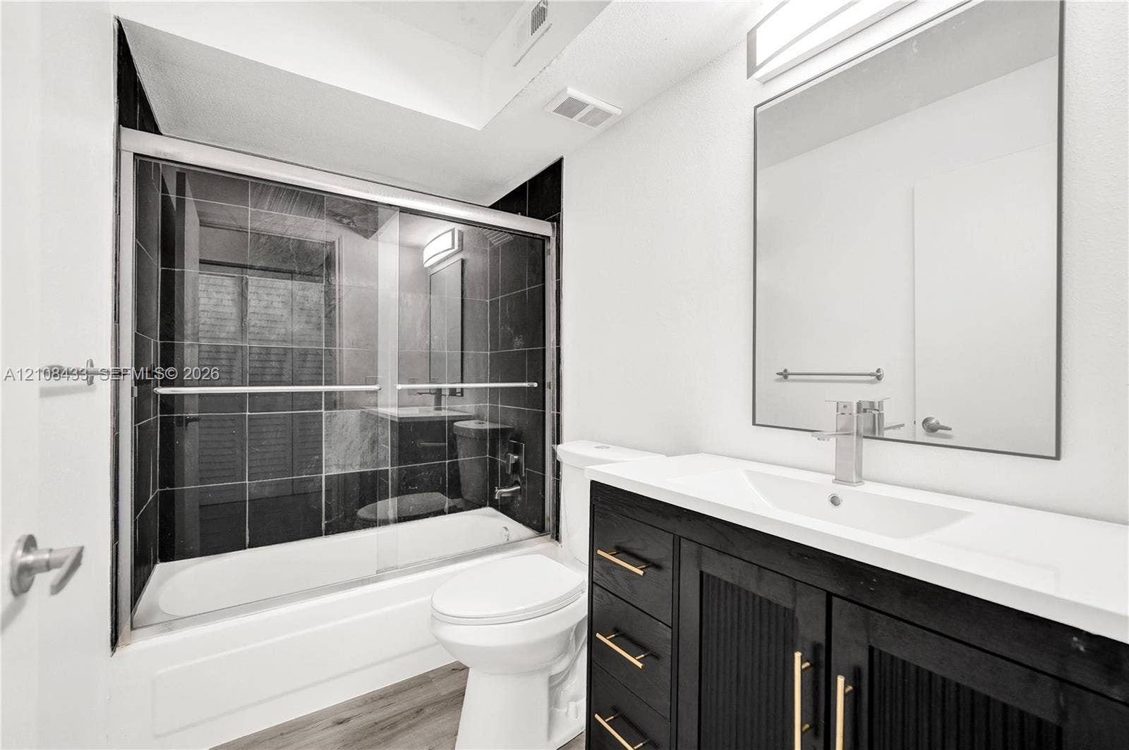 2300 NW 33rd Street, #911, Oakland Park, FL 33309 Bathroom featuring a modern vanity with a solid-surface countertop, an integrated rectangular sink, black cabinetry with gold-tone hardware, a large frameless mirror, and a shower-tub combination with dark tile surround