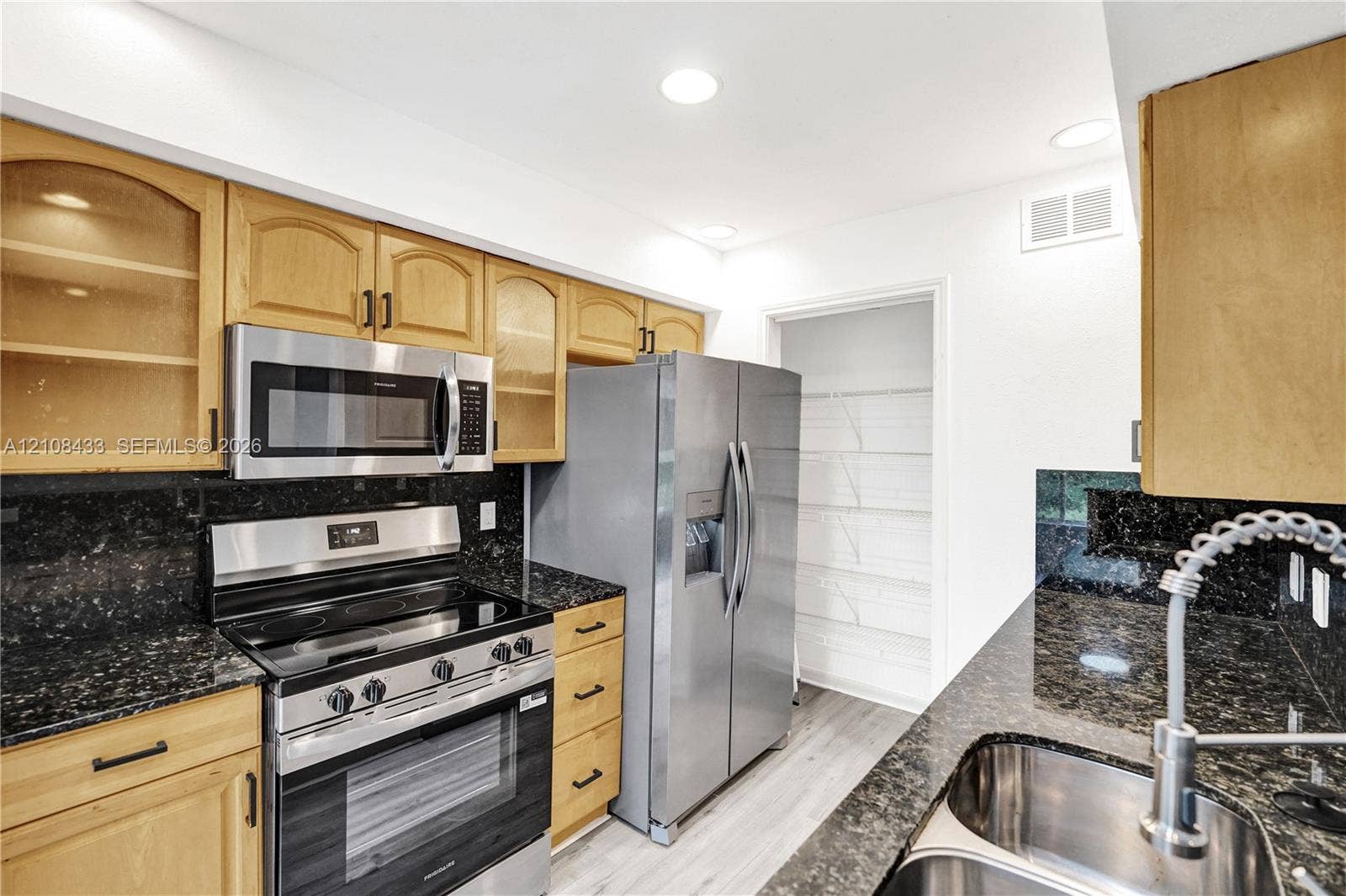 2300 NW 33rd Street, #911, Oakland Park, FL 33309 Kitchen featuring light-finish cabinetry with glass-front accents, dark stone countertops, stainless steel appliances, dual-basin stainless steel sink with pull-down faucet, and walk-in pantry