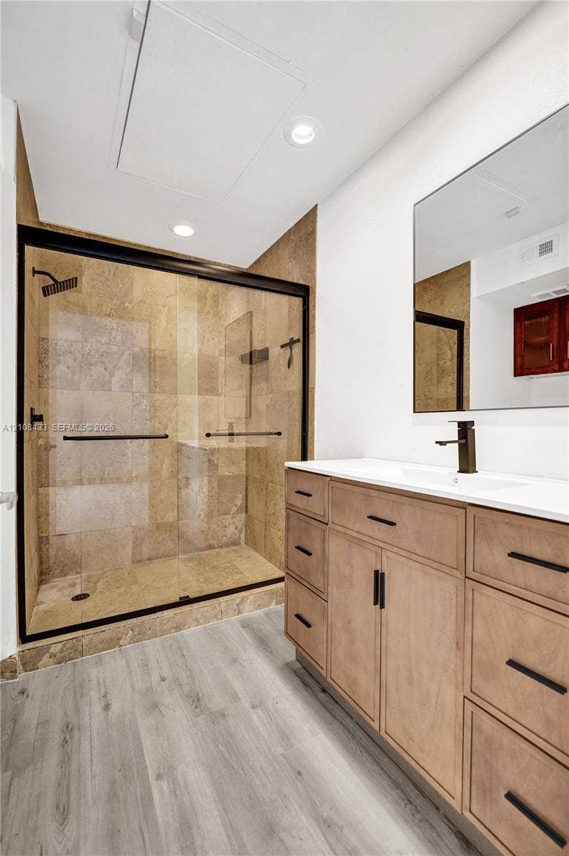 2300 NW 33rd Street, #911, Oakland Park, FL 33309 Walk-in shower with stone tile surround, glass enclosure, and matte black fixtures, paired with a wood-finish vanity, solid surface countertop, and recessed lighting