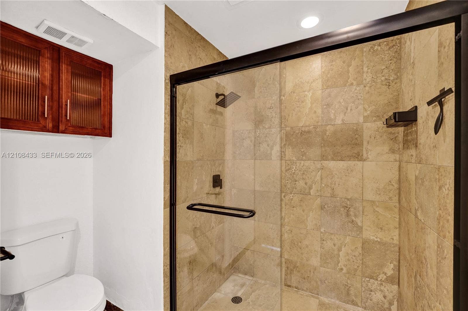 2300 NW 33rd Street, #911, Oakland Park, FL 33309 Walk-in shower with natural stone tiling, matte black frame, glass door, and overhead rainfall showerhead, complemented by wall-mounted wood-finish cabinetry