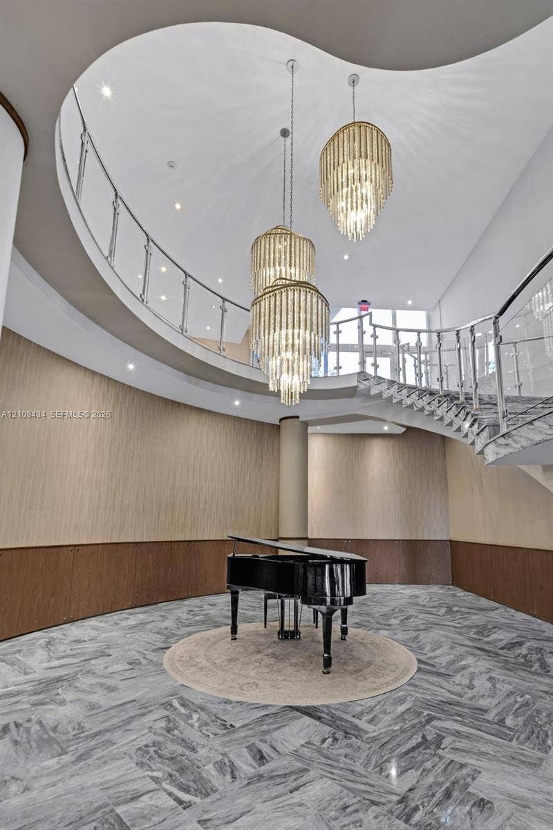 Miami Beach, FL 33140 Grand foyer featuring a sweeping curved staircase with polished metal railings, tiered crystal chandeliers, stone flooring in a herringbone pattern, and wood-paneled walls