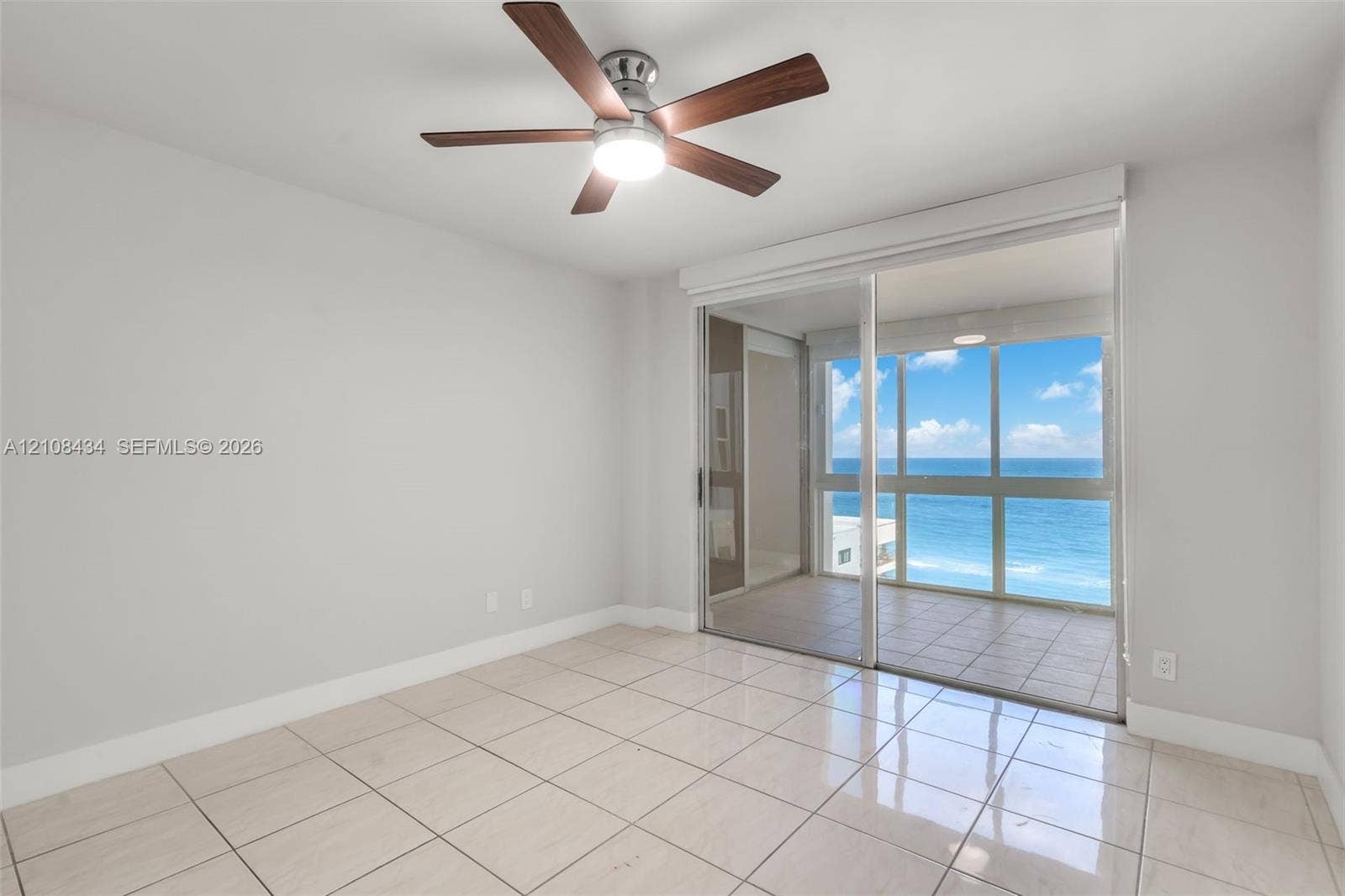 Miami Beach, FL 33140 Interior space featuring large-format ceramic tile flooring, ceiling fan with integrated lighting, and full-height sliding glass doors opening to a tiled exterior balcony with an ocean backdrop