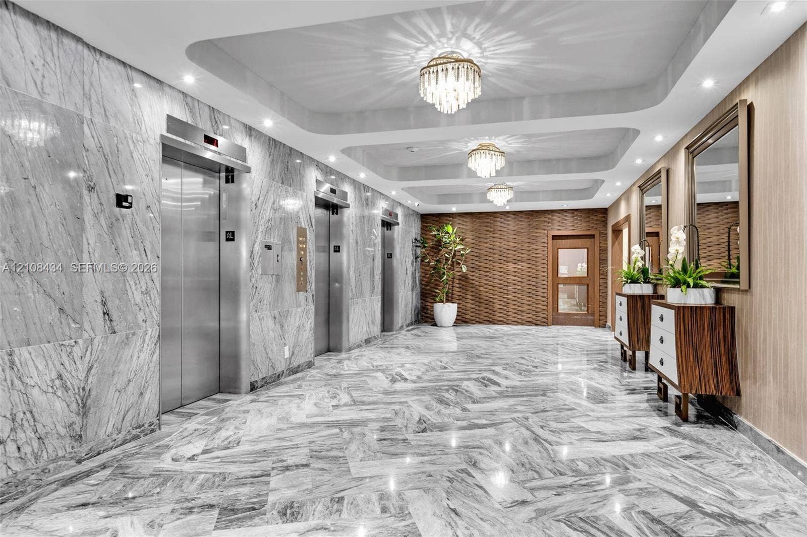 Miami Beach, FL 33140 Polished stone flooring in a herringbone pattern, full-height stone wall cladding, stainless steel elevator doors, recessed tray ceiling with multi-tiered crystal chandeliers, and textured wood-paneled accent walls