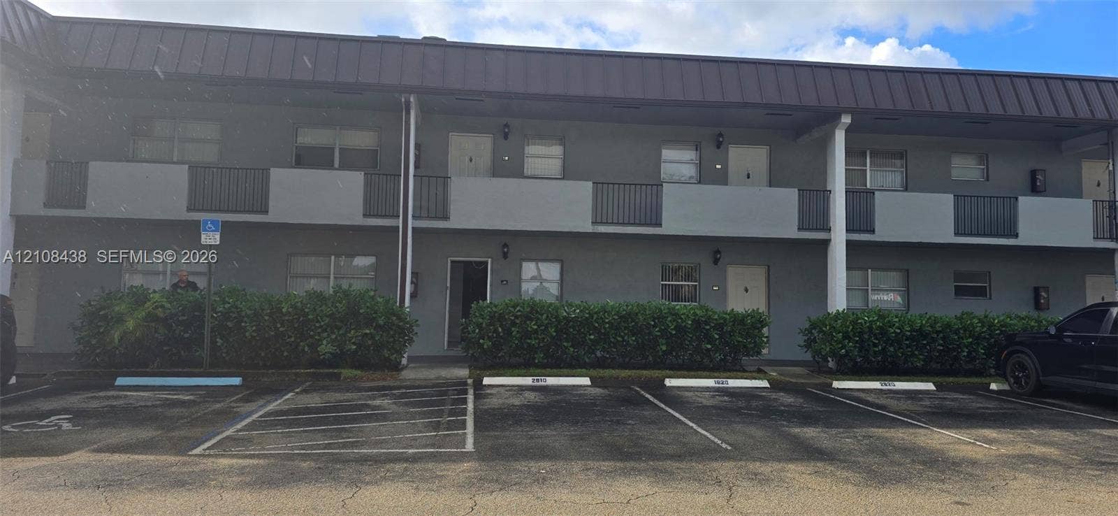 7320 NW 85th Court, #102, Tamarac, FL 33321 Two-story exterior facade featuring dark-toned metal roofing, neutral stucco siding, black metal balcony railings, and manicured foundation shrubbery